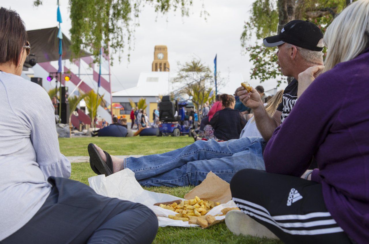 People sitting on the grass eating fish and chips at a community event, the clock tower behind.