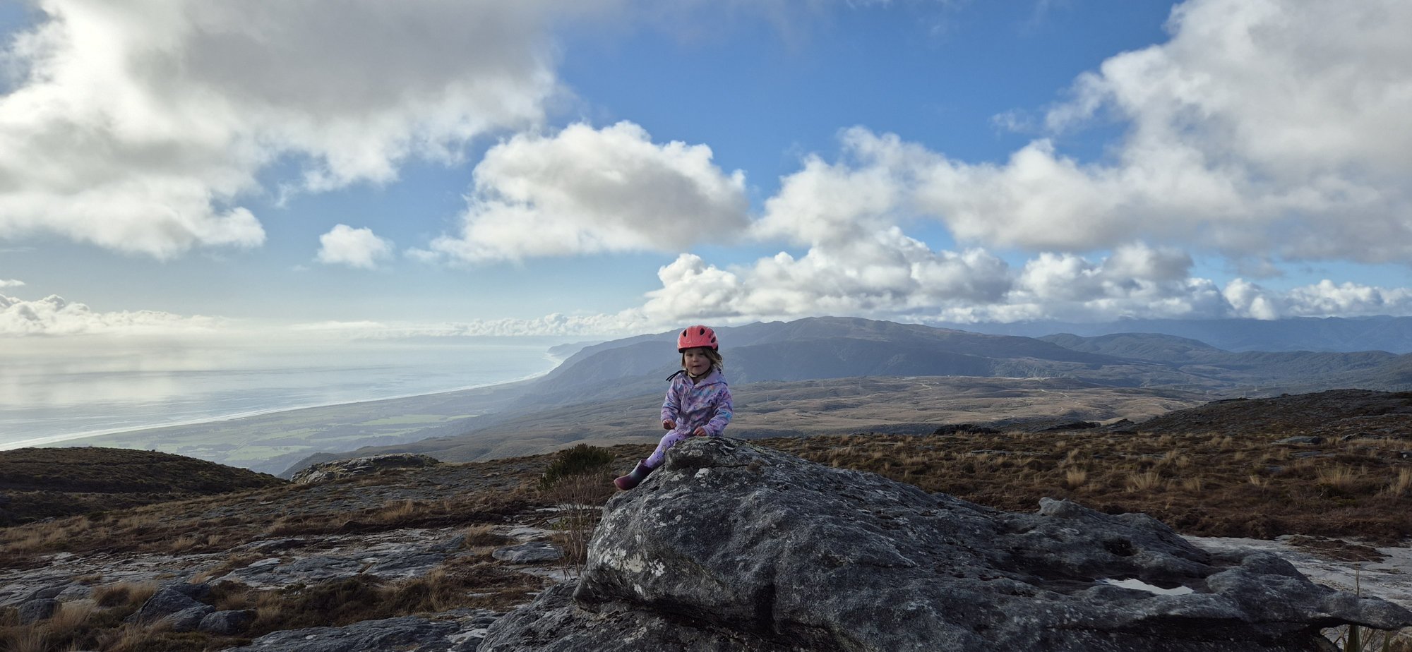 A small child in a bike helmet sitting on a rock on the plateau, with the coastline far below.