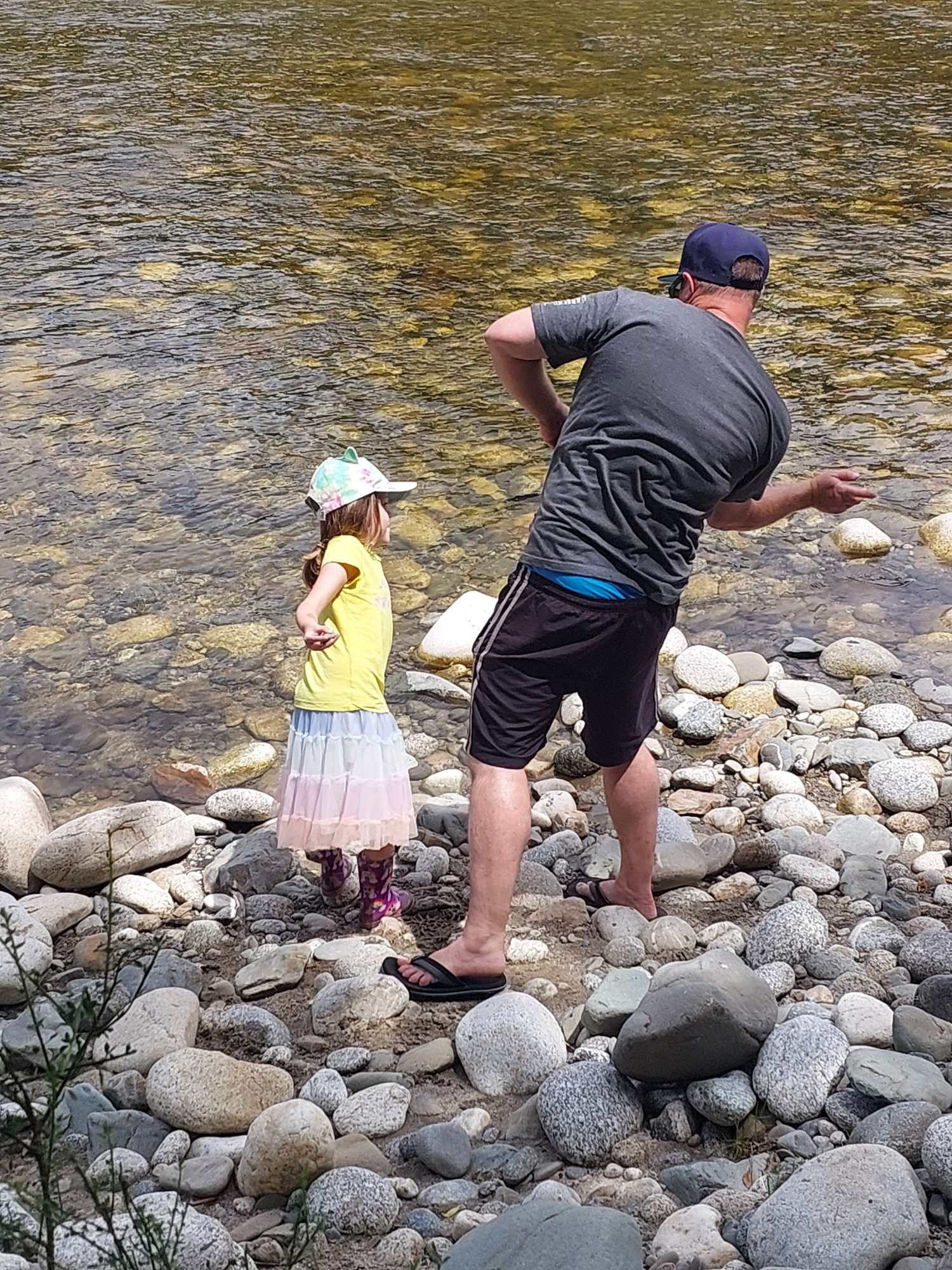 A father and young daughter skimming stones from a rocky river bank.