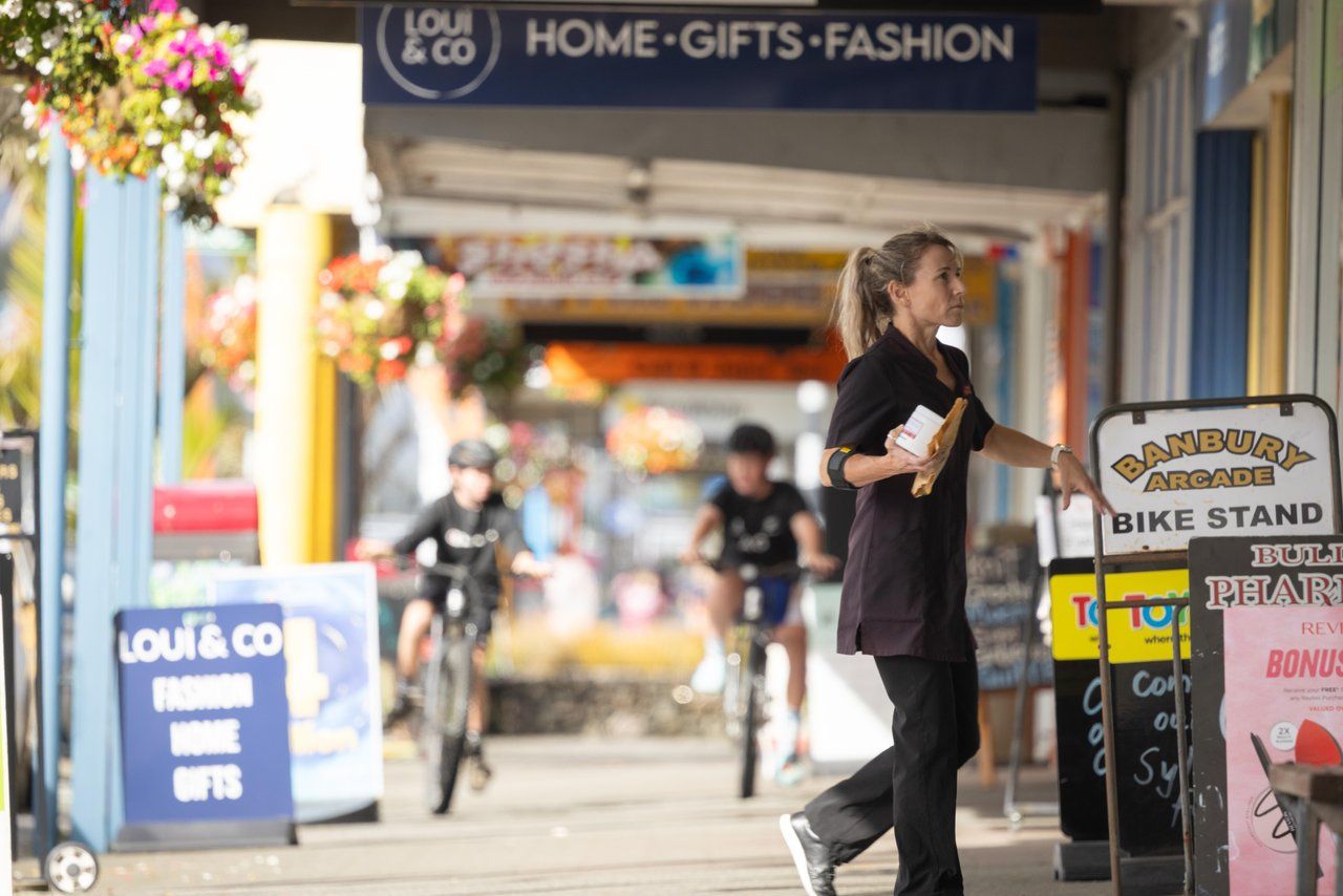 A shop worker stepping out onto the street with a coffee, children cycling past.